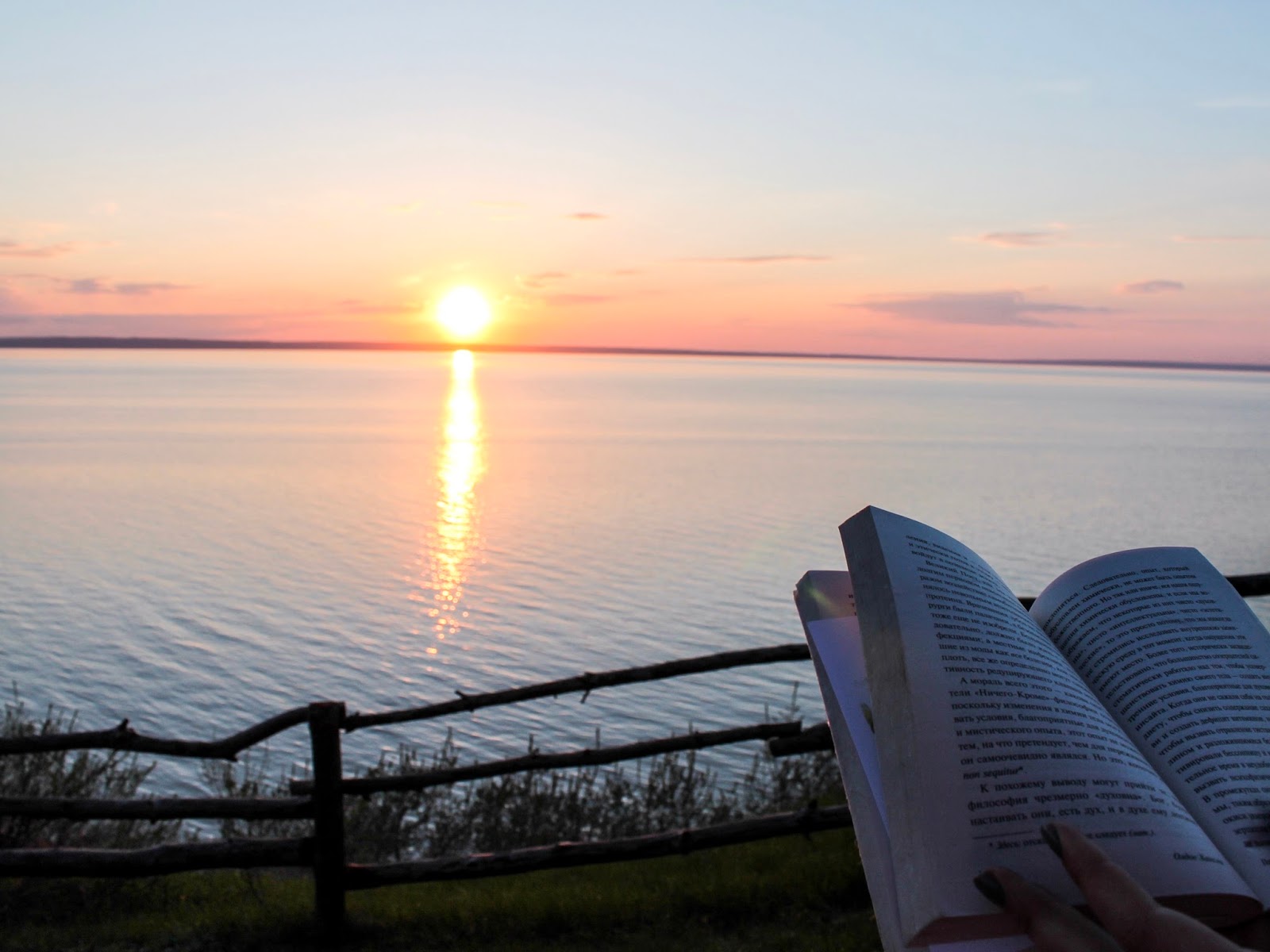 Человек читает книгу на закате у воды, вид с деревянным забором