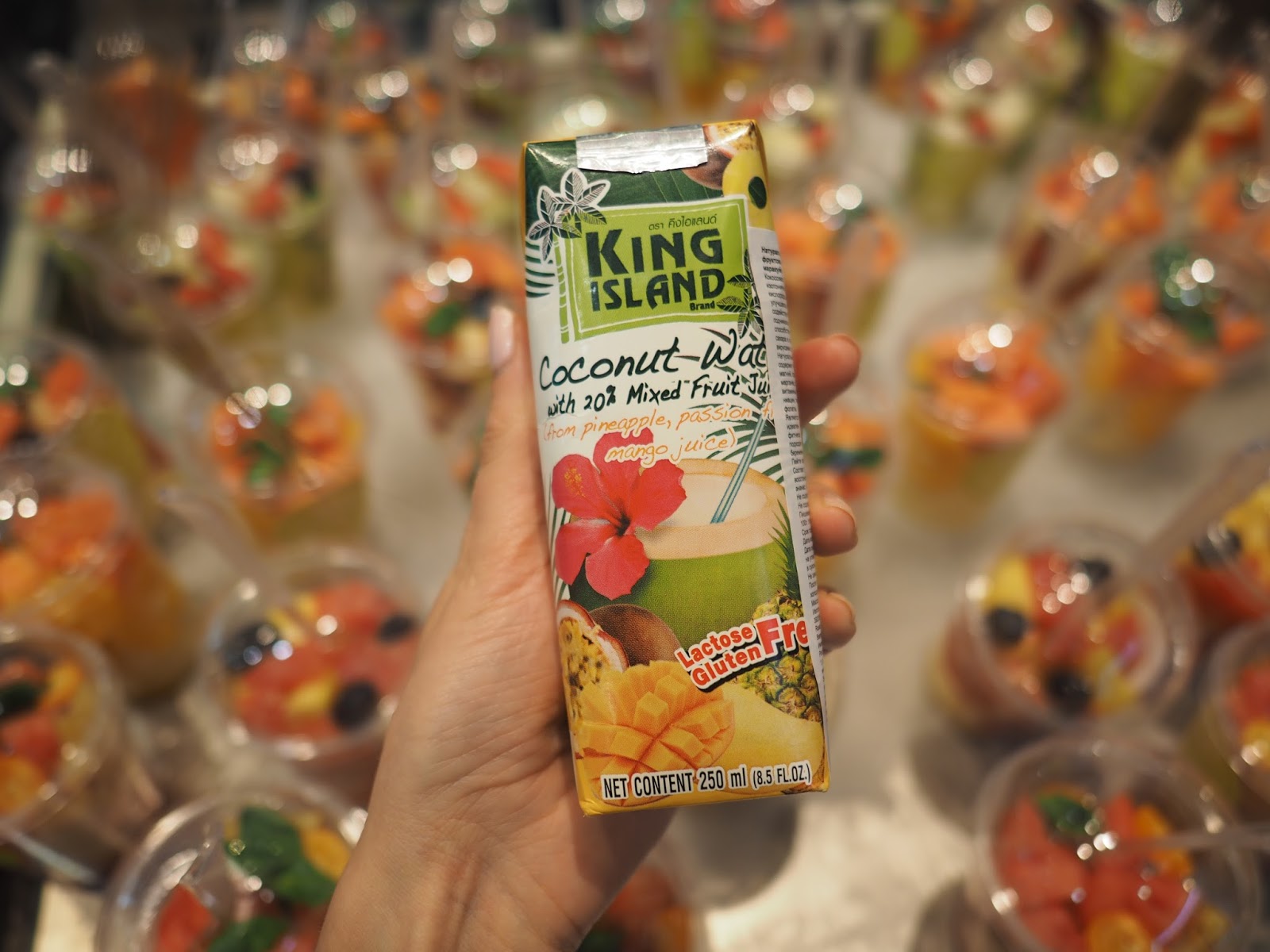 Упаковка кокосовой воды King Island с манго и маракуйей на фоне стаканов с фруктовым салатом