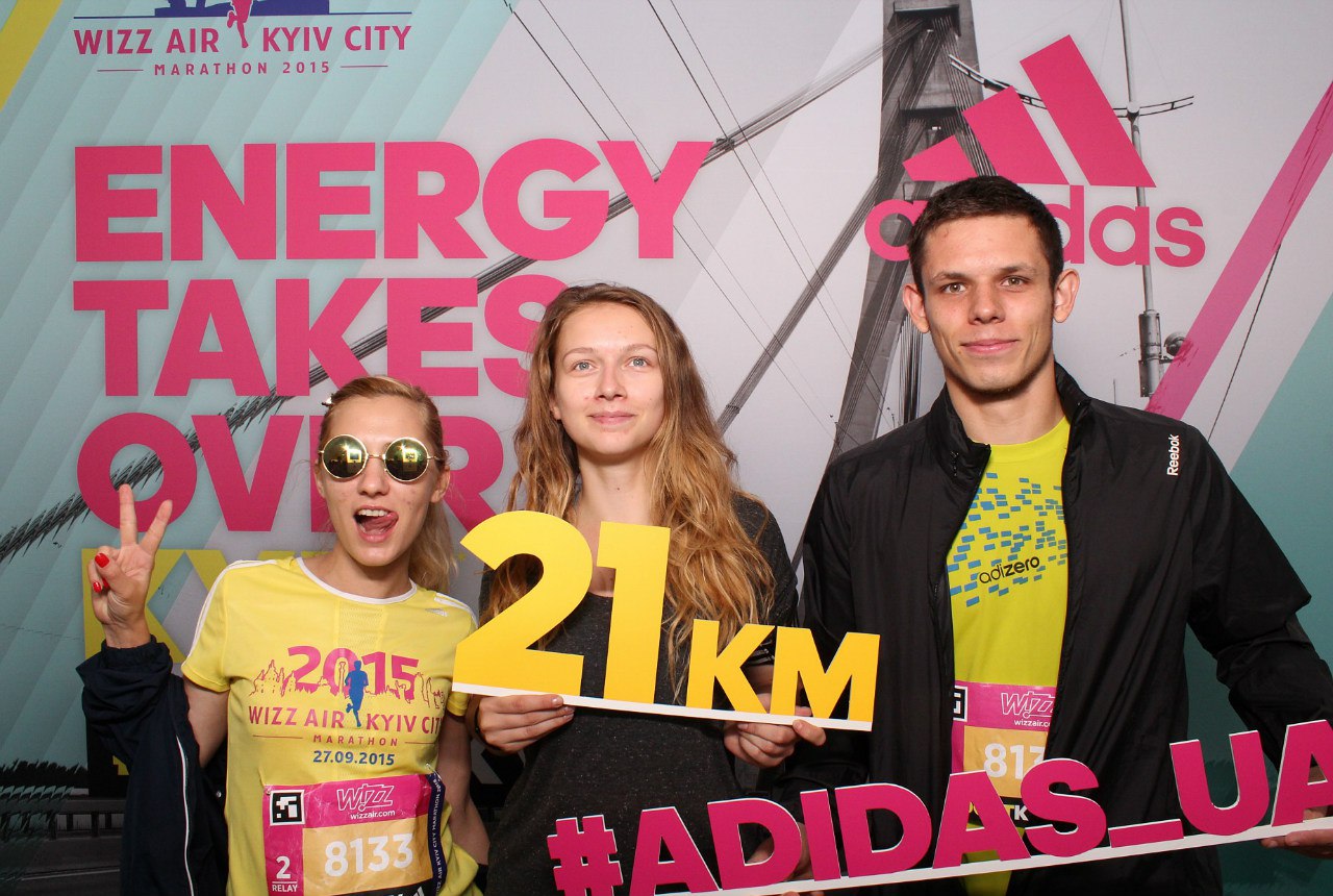 Трое участников марафона Wizz Air Kyiv City держат табличку 21 км на фоне рекламного стенда adidas